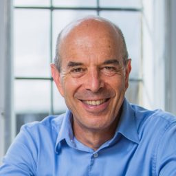 Smiling man with a bald head wearing a blue shirt sits in front of a window.