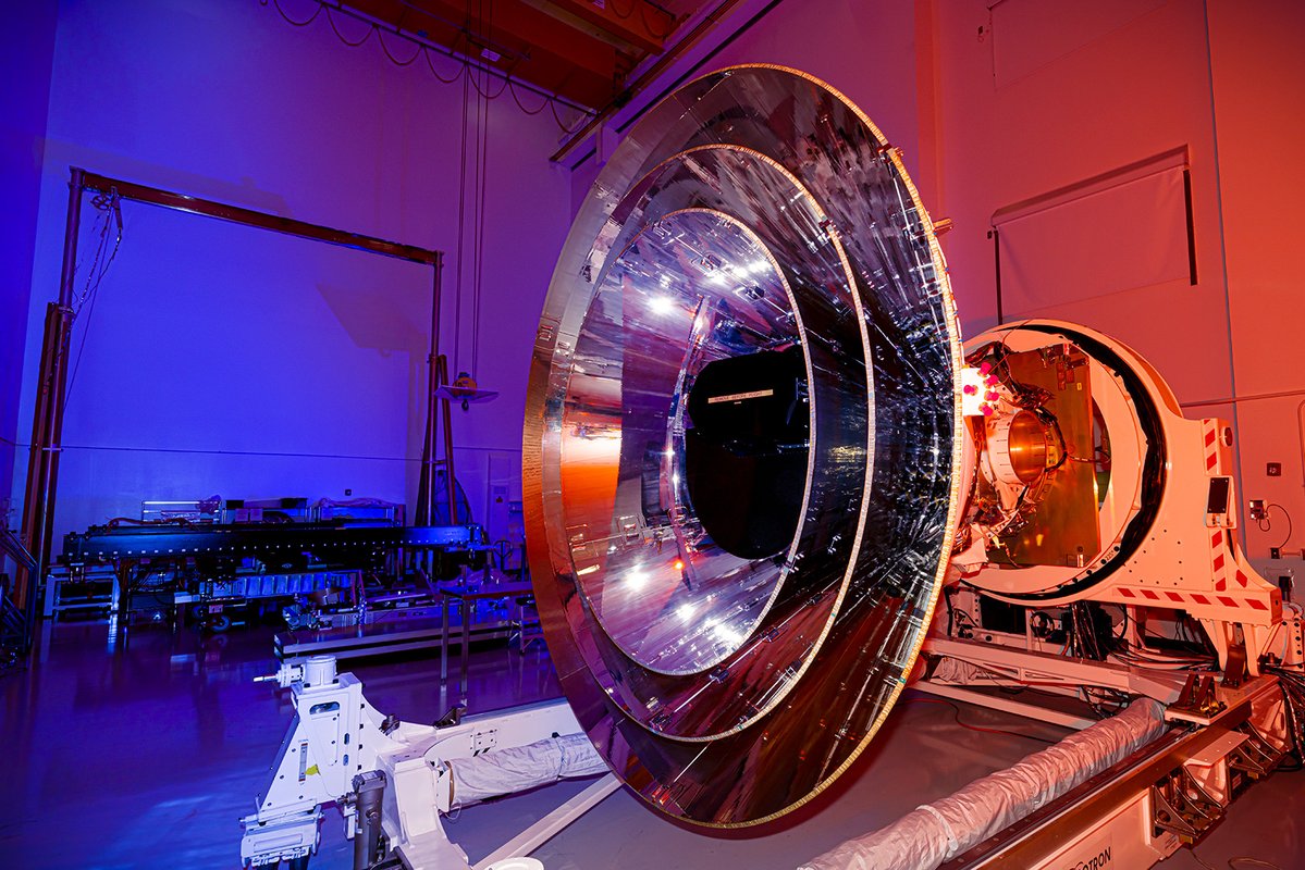 A large, reflective satellite dish inside a dimly lit laboratory with purple and orange lighting, surrounded by equipment and machinery, is poised to map galaxies, illuminating mysteries that even the CMB can't teach us.