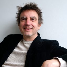A man with tousled hair, wearing a black jacket and light sweater, sits against a white wall, smiling at the camera.