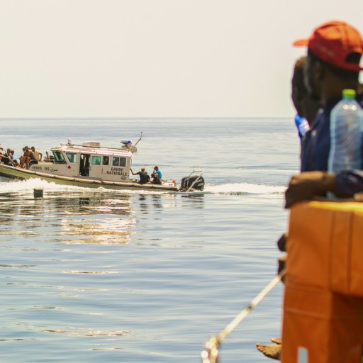 A group of people on a boat watch as a patrol vessel approaches on calm, open water under a clear sky, reflecting ongoing migration statistics in the region.