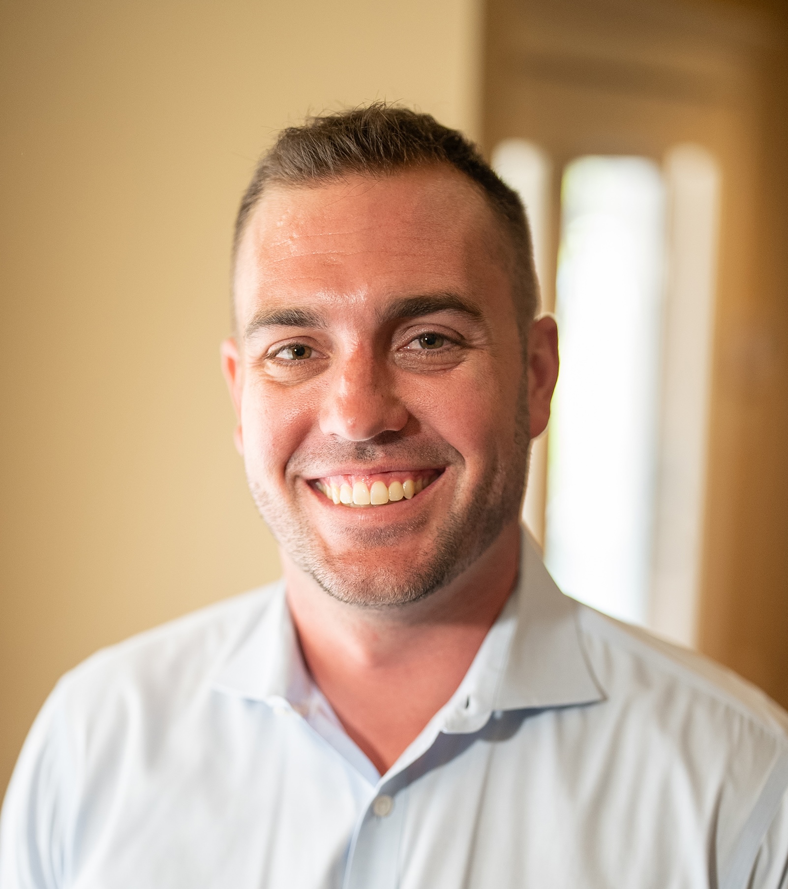 Smiling man in a light blue shirt stands indoors with a blurred doorway in the background.