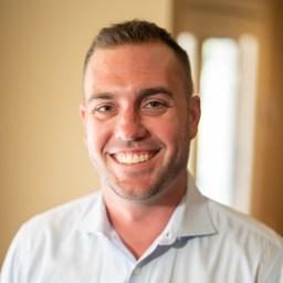 Smiling man in a light blue shirt stands indoors with a blurred doorway in the background.