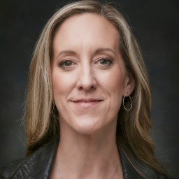 A person with long blonde hair and a leather jacket smiles slightly against a dark background.