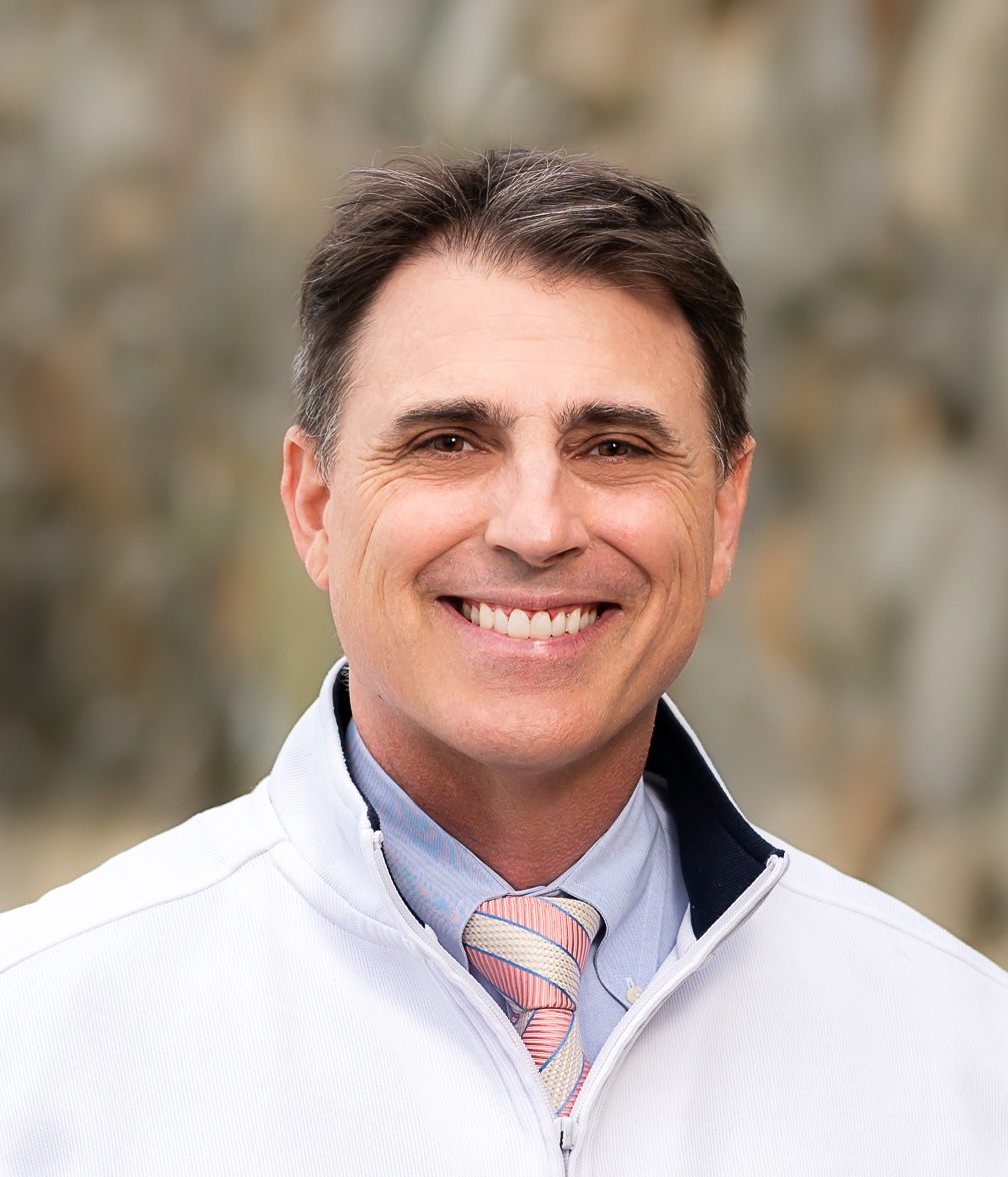 Smiling man with short brown hair, wearing a light jacket, blue shirt, and pink-striped tie, stands against a blurred background.