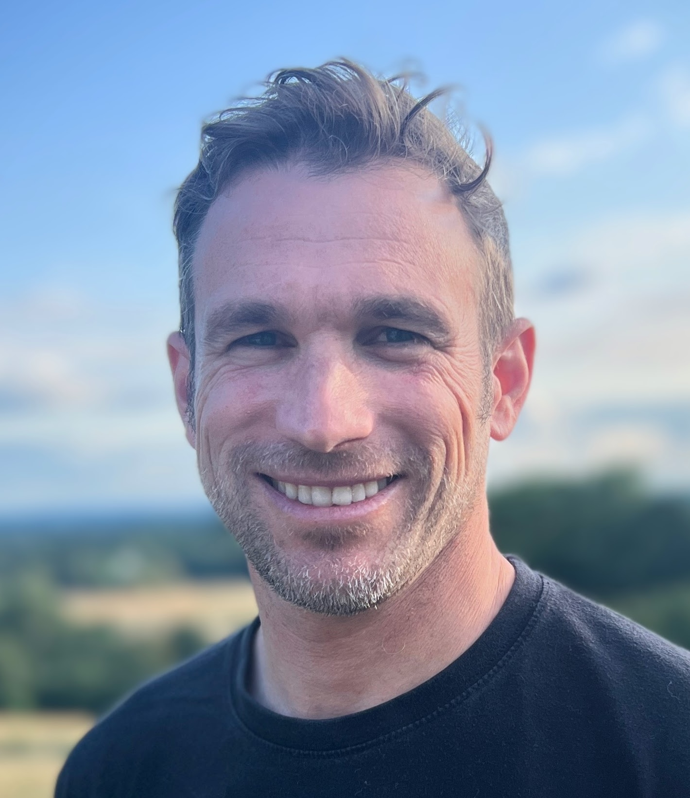 A man with short hair and a beard smiles at the camera, standing outdoors with a blurred landscape and blue sky in the background.