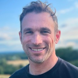 A man with short hair and a beard smiles at the camera, standing outdoors with a blurred landscape and blue sky in the background.