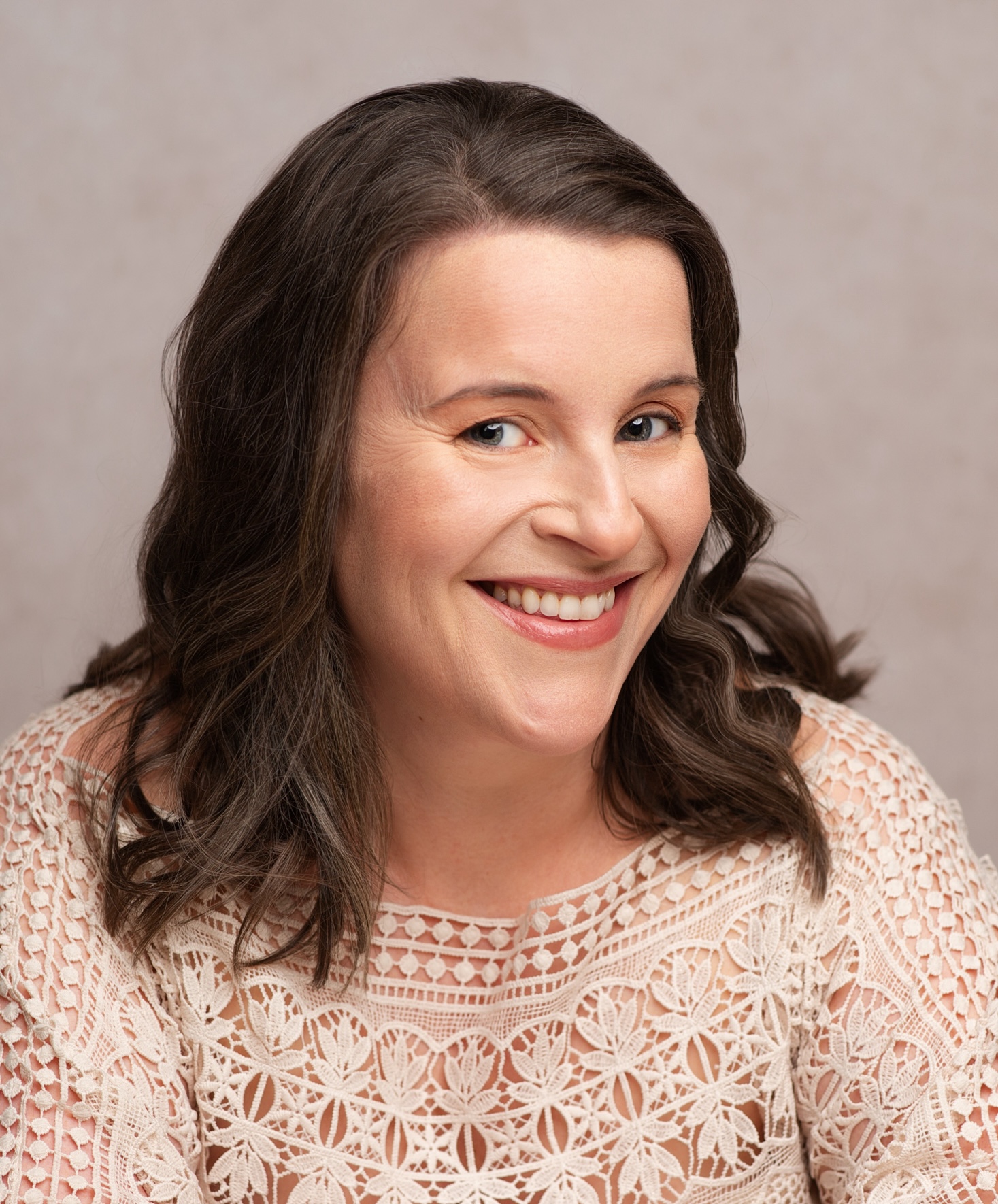 A smiling person with wavy brown hair wearing a lace top poses against a neutral background.