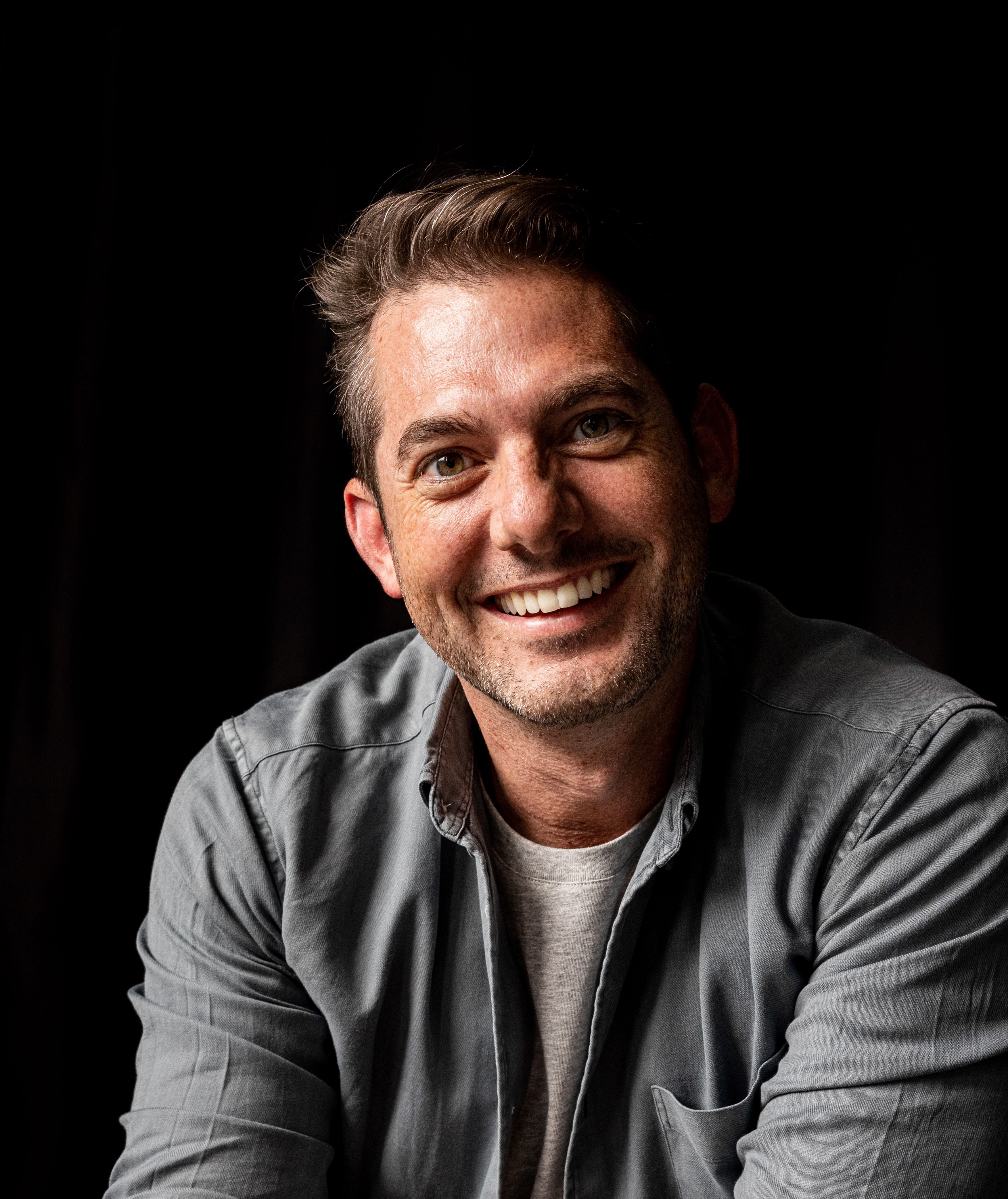 Man with short hair and a stubbly beard, smiling while wearing a gray shirt over a white tee, against a dark background.