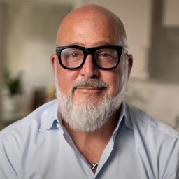 A person with a bald head, white beard, and black glasses is wearing a light blue shirt, standing in a softly lit kitchen.