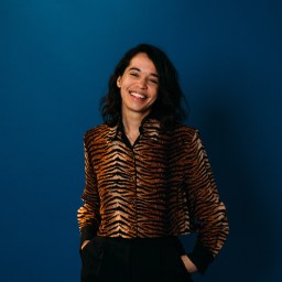 A person with dark hair smiles, wearing a tiger-striped shirt against a dark blue background.
