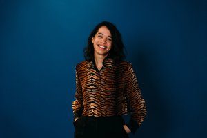 A person with dark hair smiles, wearing a tiger-striped shirt against a dark blue background.