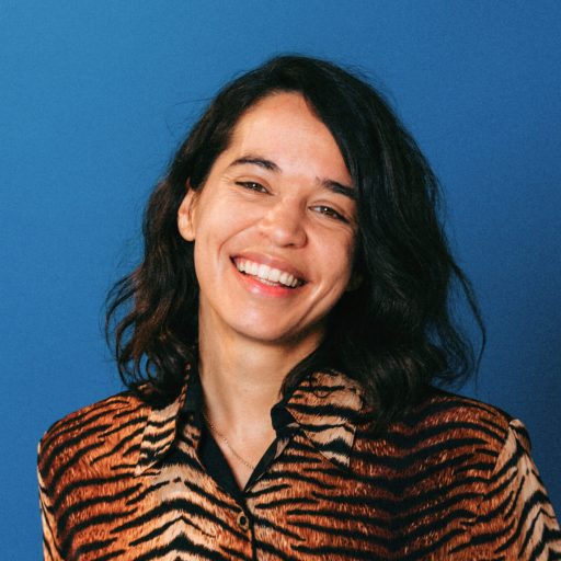 A person with long dark hair beams with creativity, wearing a tiger print jacket, set against a vibrant blue background.