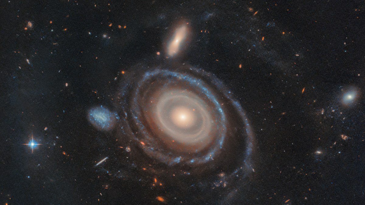 A spiral galaxy with a luminous core, surrounded by swirling arms and smaller galaxies, forms a mesmerizing bullseye ring galaxy, set against a backdrop of stars in space.