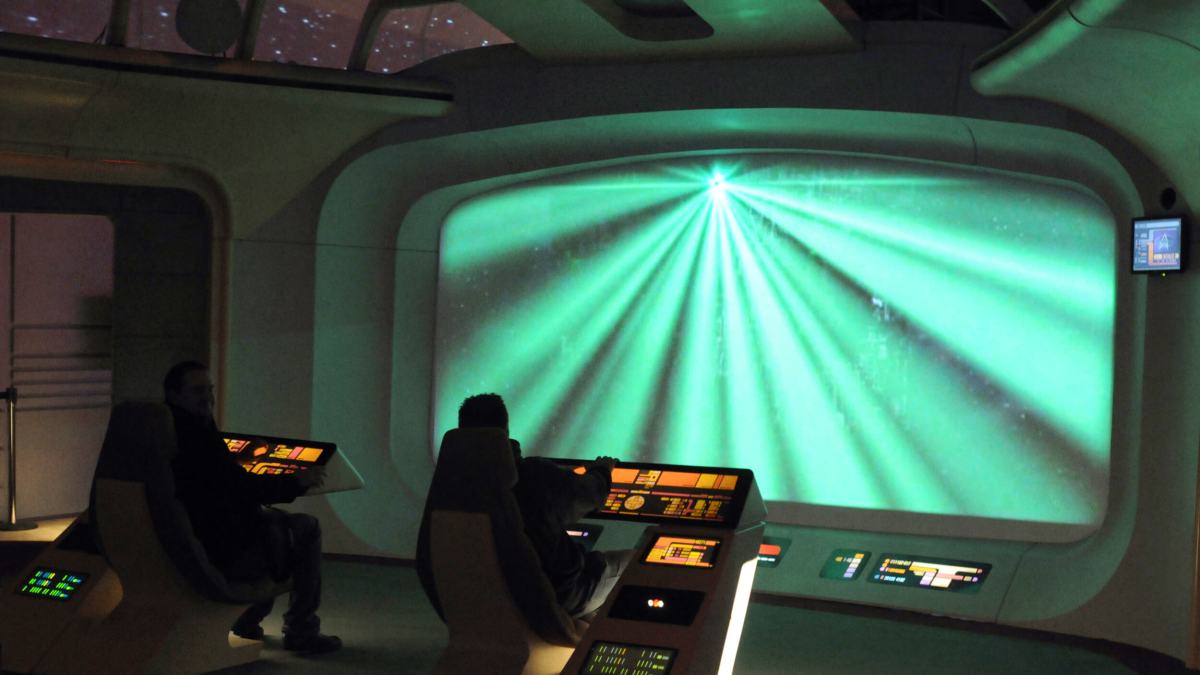 In a futuristic control room, two people sit at advanced control panels, utilizing AI technology as they face a large screen displaying vibrant green light beams.
