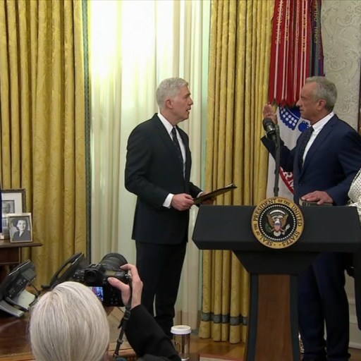Group of people in a formal setting, with a man holding a large book, others standing nearby, and photographers capturing the scene. There is a large portrait and flags in the background.