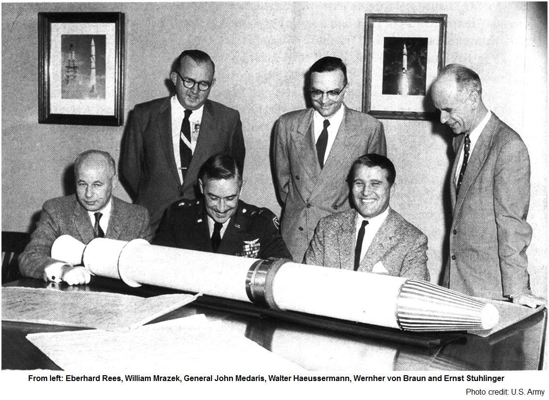 Six men in suits and one in military uniform examine a rocket model and blueprints on a table, showcasing their commitment to fundamental research that drives innovation and invites significant investment.