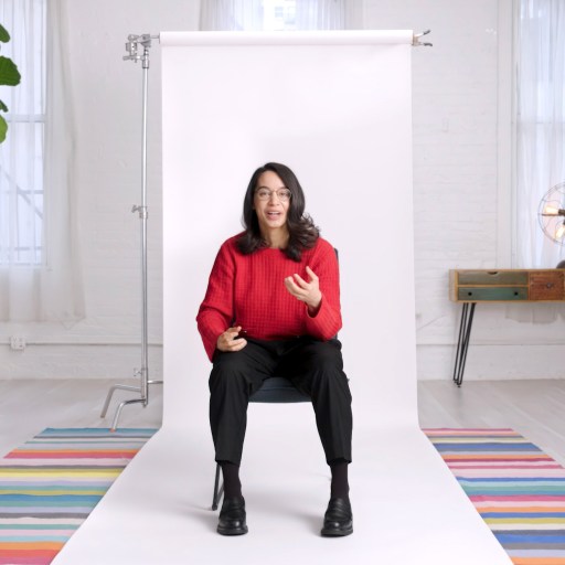 Person in a red sweater sitting on a stool in a bright studio with white walls and colorful striped rugs.