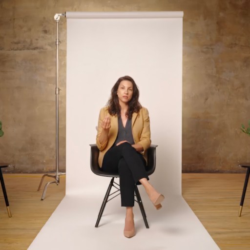 A person sitting on a chair in a studio setting with a white backdrop and two small tables on either side, each holding a small plant.