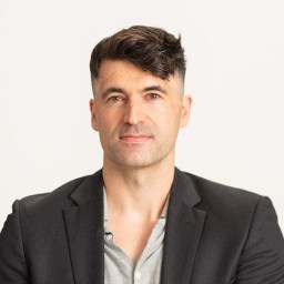 Man with short dark hair wearing a black blazer and gray shirt against a light background.