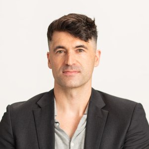 Man with short dark hair wearing a black blazer and gray shirt against a light background.