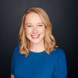 Smiling person with shoulder-length blonde hair, wearing a blue top against a dark background.