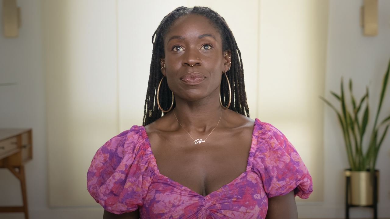 Person wearing a purple floral top and hoop earrings, looking upwards. A plant and a wooden table are in the background.