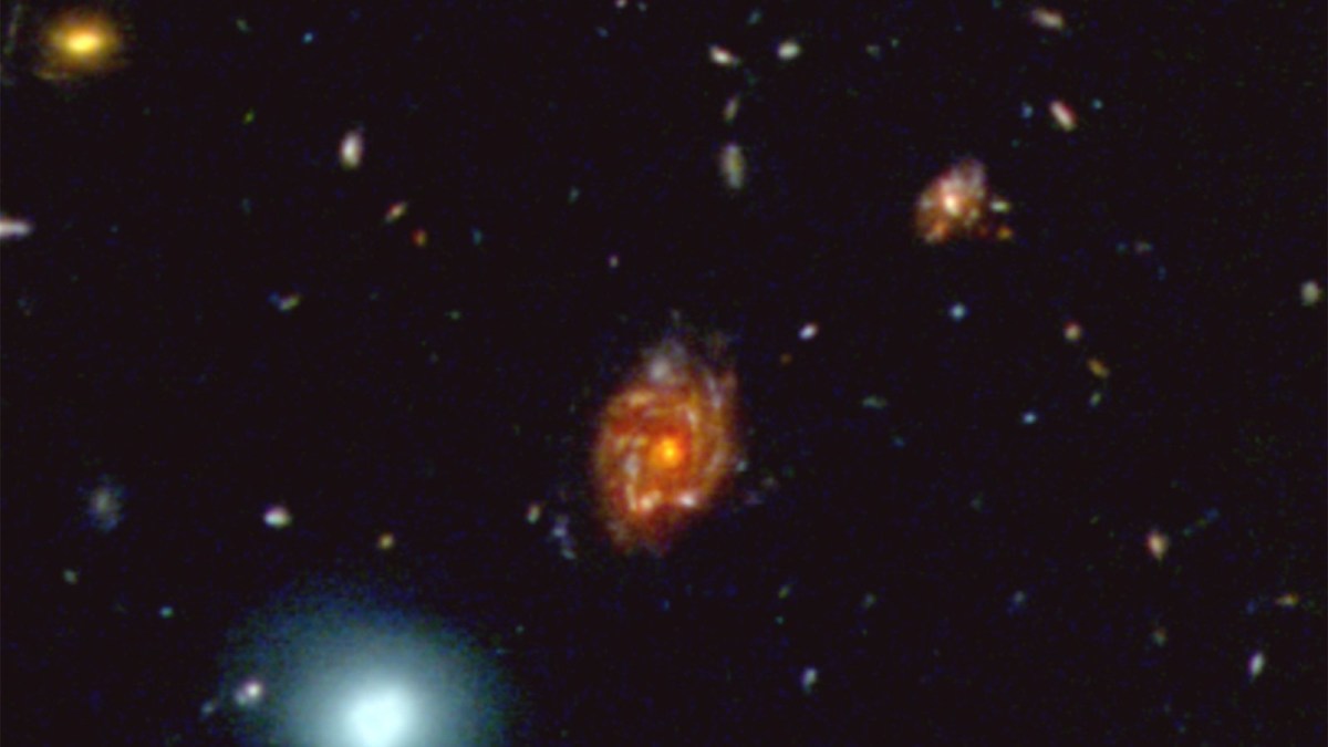 A colorful, spinning galaxy with a bright orange core, existing for 12 billion years, is surrounded by smaller galaxies and star clusters against a black space background.