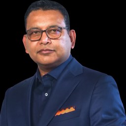 A man wearing glasses and a blue suit with an orange pocket square against a black background.