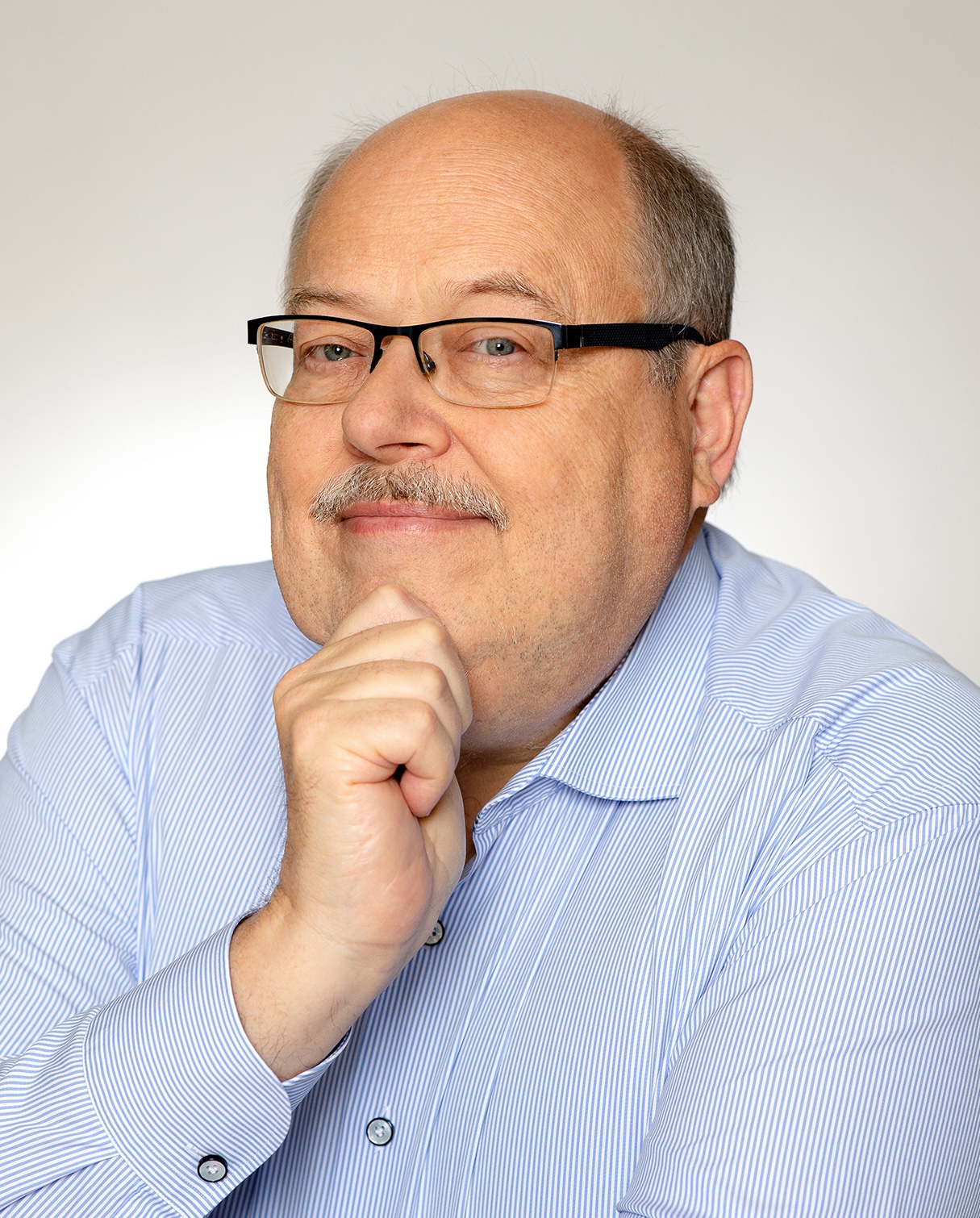 A man wearing glasses and a light blue shirt with vertical stripes, resting his chin on his hand, poses against a plain background.