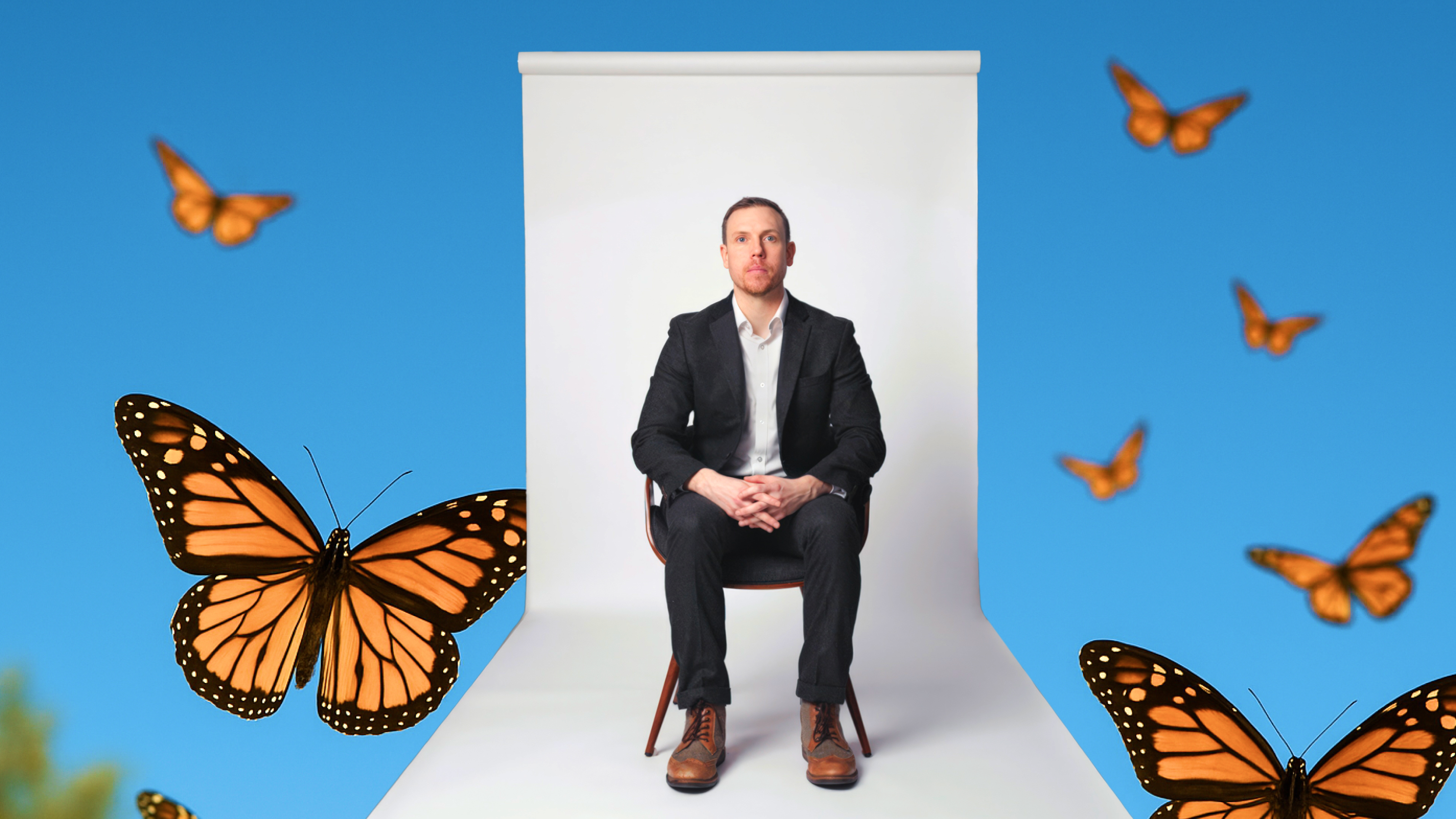A man in a black suit sits on a chair against a white backdrop, surrounded by several monarch butterflies on a bright blue background.