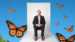A man in a black suit sits on a chair against a white backdrop, surrounded by several monarch butterflies on a bright blue background.