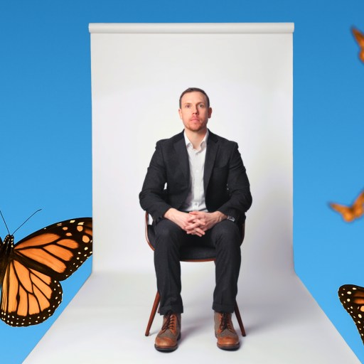 A man in a black suit sits on a chair against a white backdrop, surrounded by several monarch butterflies on a bright blue background.