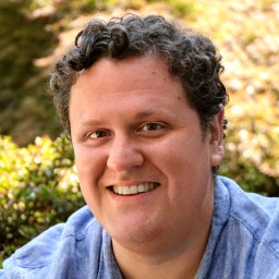 Smiling person with curly hair wearing a blue shirt in an outdoor setting with greenery.
