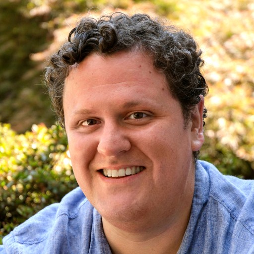 Smiling person with curly hair wearing a blue shirt in an outdoor setting with greenery.