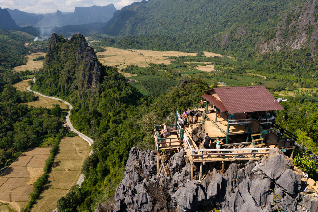 A wooden viewing platform with people on it sits atop a rocky hill, overlooking green valleys, mountains, and fields under a partly cloudy sky.