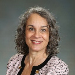 A woman with curly gray hair smiles while wearing a patterned jacket against a gray backdrop.