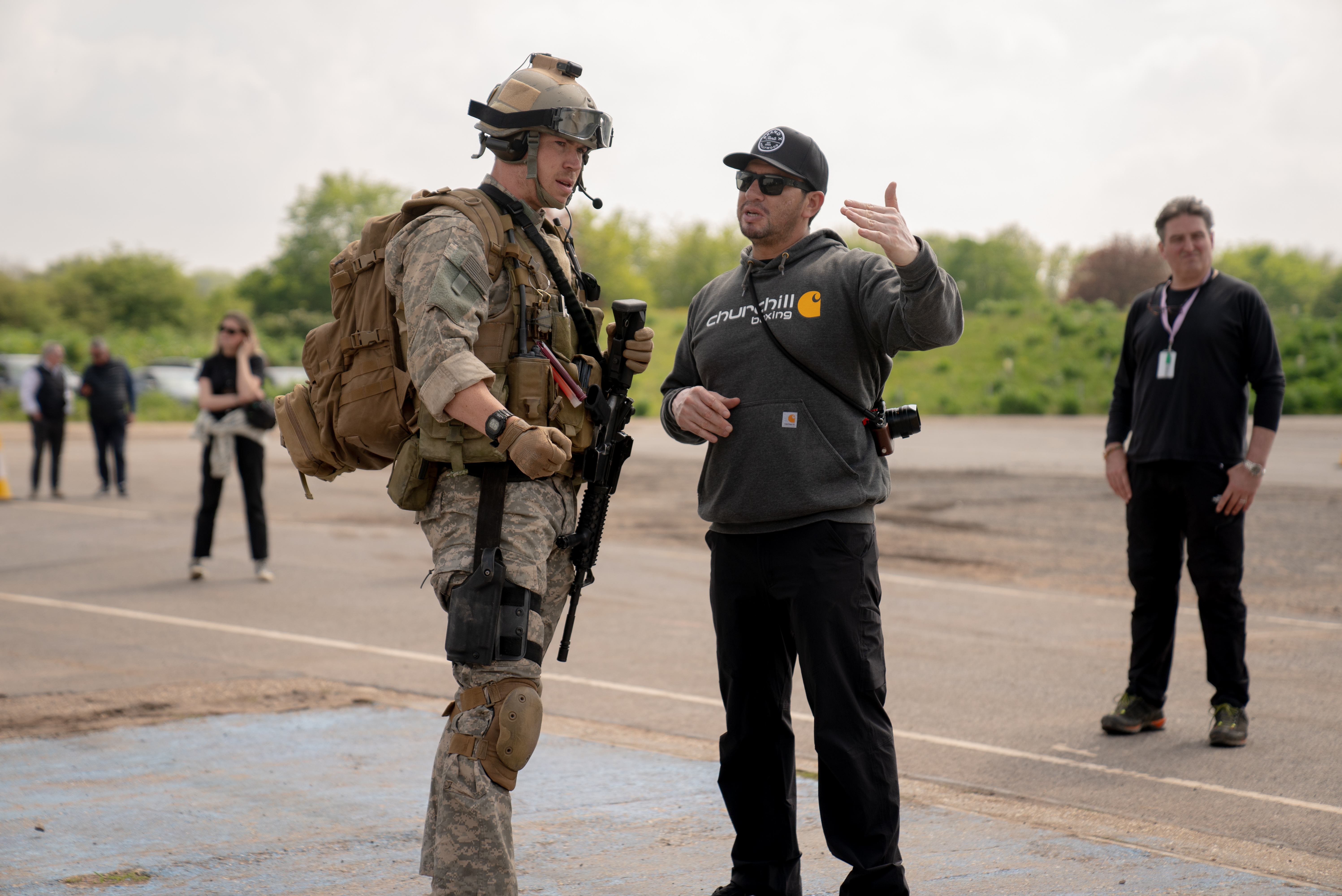 A person in military gear holding a rifle listens to someone in casual clothing who is gesturing, evoking the tense atmosphere found in an Alex Garland film, with others standing in the background outdoors.