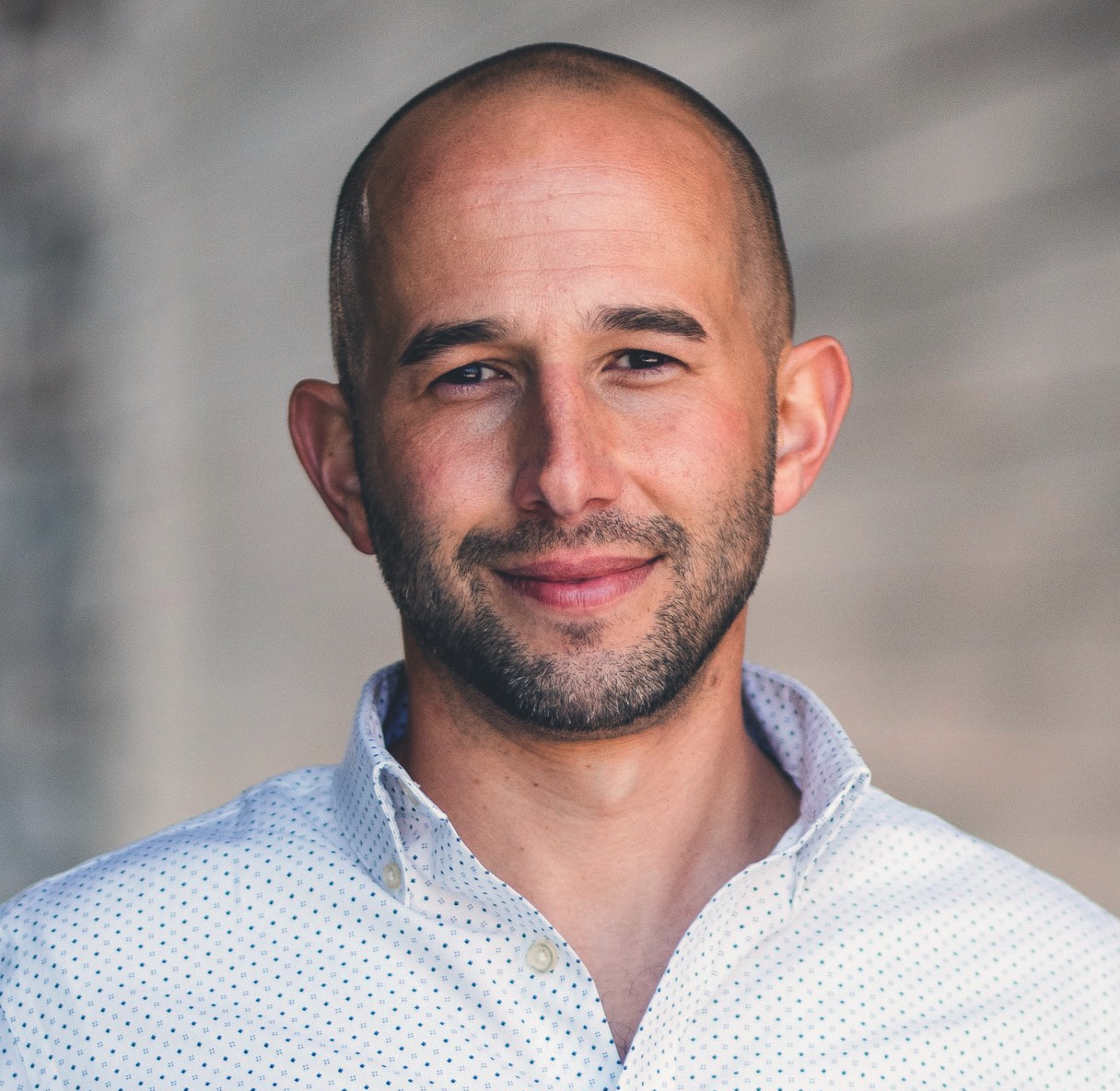 A bald man with a trimmed beard wearing a white, patterned button-up shirt stands in front of a blurred, neutral-toned background.