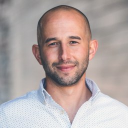 A bald man with a trimmed beard wearing a white, patterned button-up shirt stands in front of a blurred, neutral-toned background.