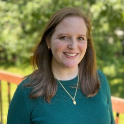 Woman with long brown hair wearing a green sweater and gold necklace smiles at the camera, standing outdoors with green trees in the background.