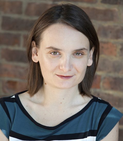 A woman with straight brown hair and light skin wearing a blue and black striped top stands in front of a brick wall, looking directly at the camera.