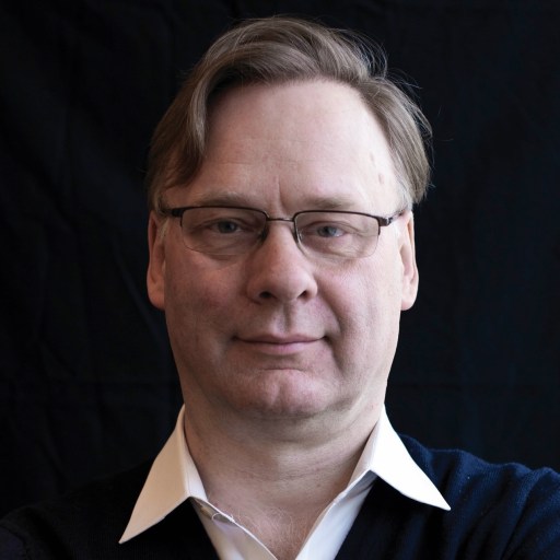 A person wearing glasses and a dark sweater over a white shirt against a dark background.