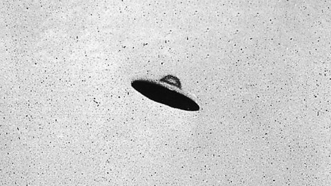 Black-and-white photo showing a disc-shaped object with a shadowed underside, floating in a blank sky—an iconic image from the UFO craze.