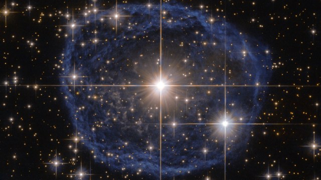 Bright star at the center of a blue nebula radiates mass energy, surrounded by numerous smaller stars, with diffraction spikes visible from the brightest stars.