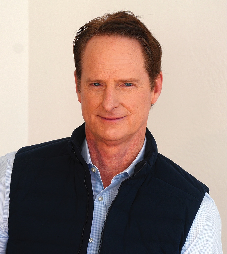 A person with short brown hair is smiling while wearing a light blue shirt and dark vest, standing against a light background.