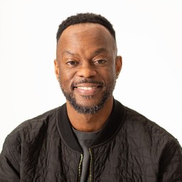 A person with a beard and short hair smiles at the camera, wearing a black quilted jacket and earrings, against a plain white background.
