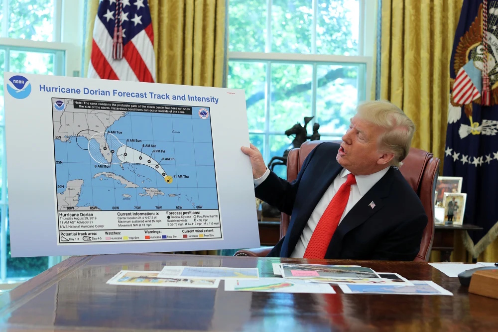 A man in a suit sits at a desk in an office, holding up a large weather map to stress the importance of truth when sharing the forecast track and intensity of Hurricane Dorian.
