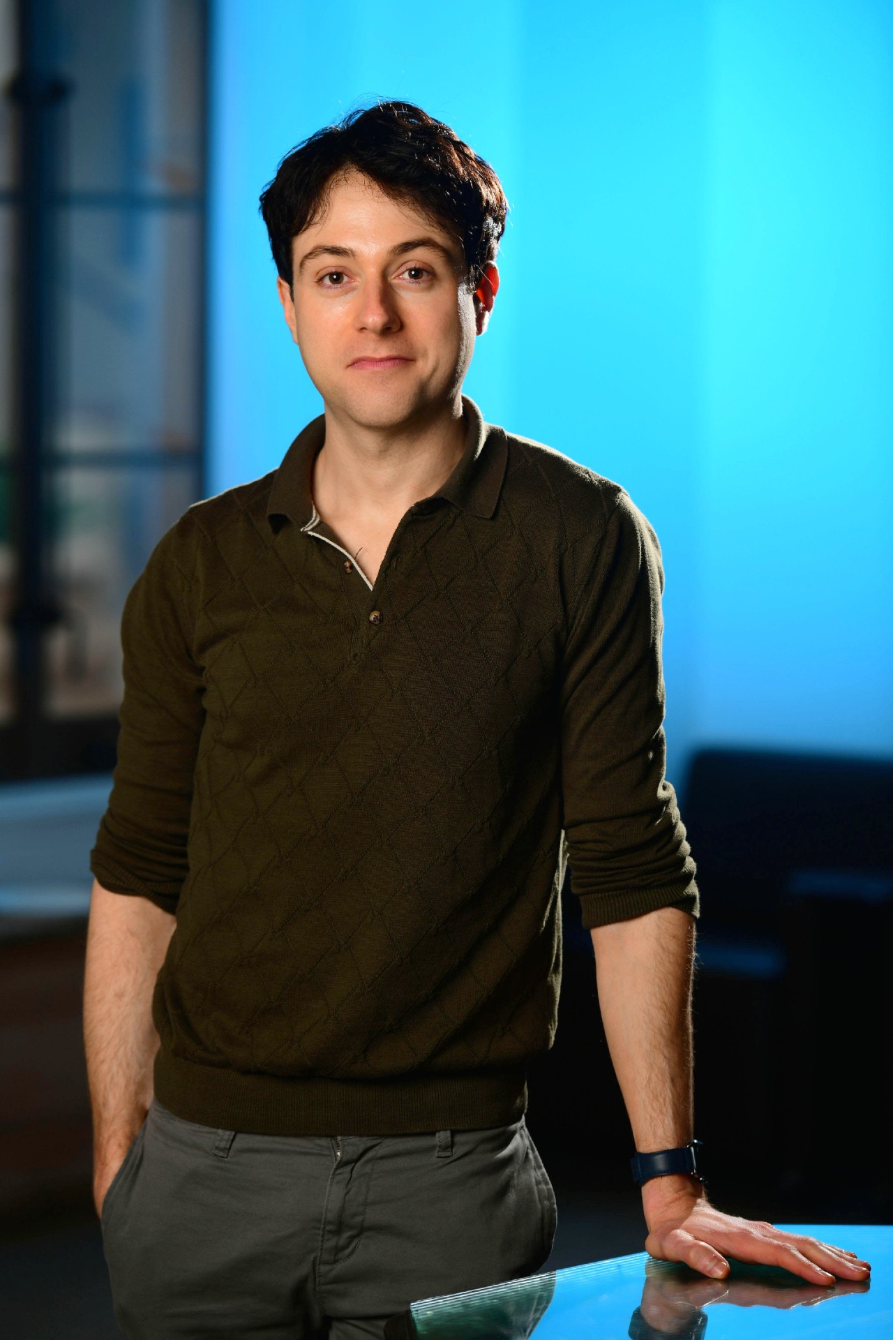 A man with short dark hair, wearing a dark green sweater and gray pants, stands indoors with one hand resting on a glass surface. The background features a blue gradient and a window.