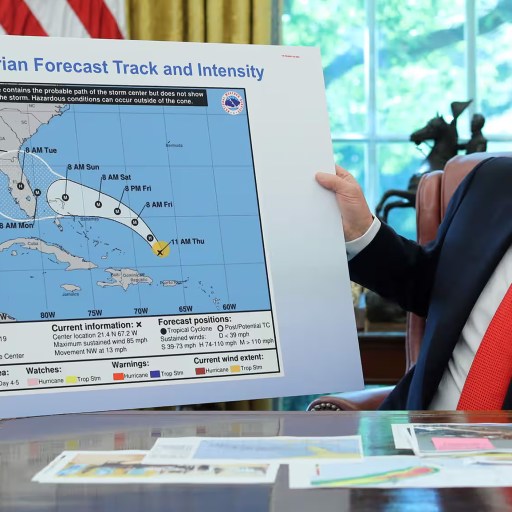 A man in a suit holds up a Hurricane Dorian forecast map in an office, tracing the storm’s projected path and intensity over several southeastern U.S. states and the Bahamas—echoing the urgent clarity of a 1938 science manifesto defending democracy.