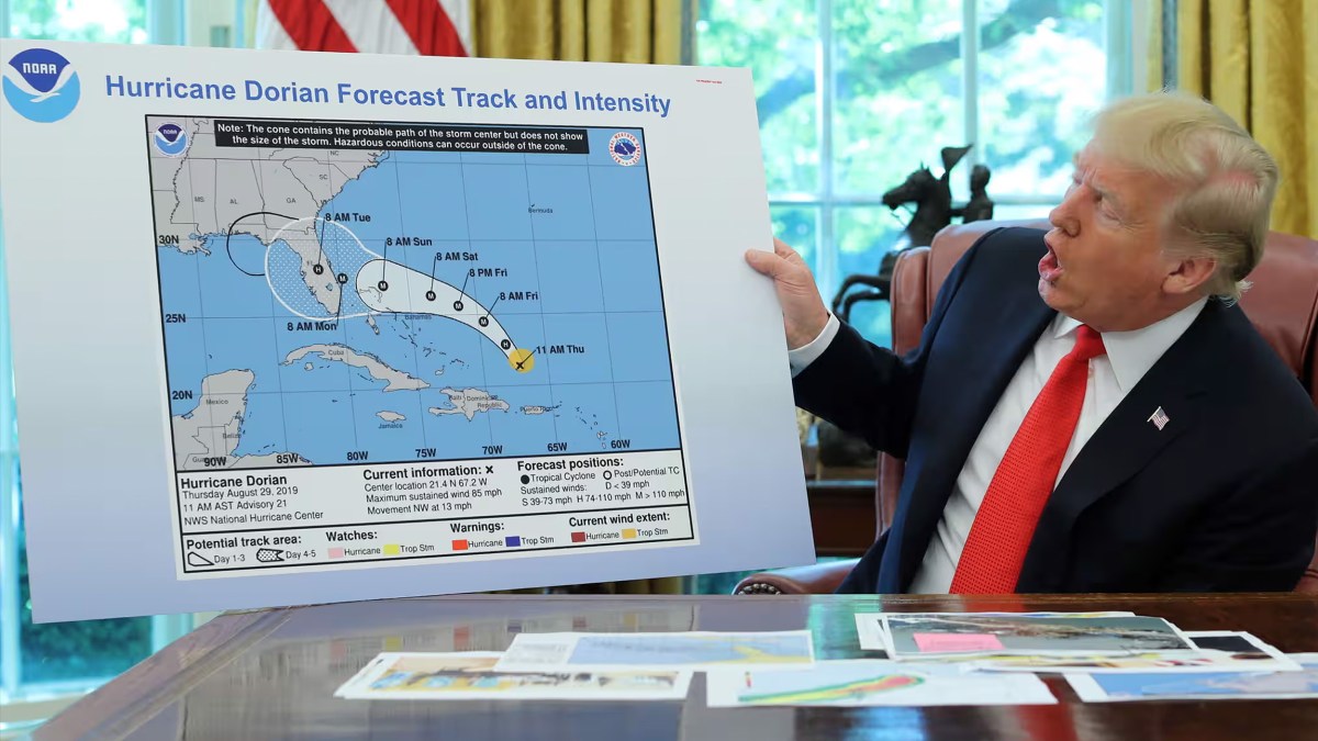 A man in a suit holds up a Hurricane Dorian forecast map in an office, tracing the storm’s projected path and intensity over several southeastern U.S. states and the Bahamas—echoing the urgent clarity of a 1938 science manifesto defending democracy.
